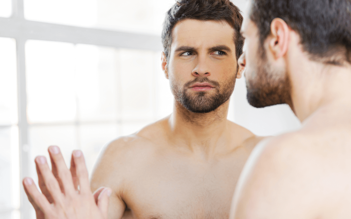 Confidence beim Sex - Symbolbild: Ein Mann mit nacktem Oberkörper schaut kritisch in den Spiegel. Sexual Confidence – symbolic image: a man with a naked torso looks critically in the mirror.