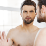 Confidence beim Sex - Symbolbild: Ein Mann mit nacktem Oberkörper schaut kritisch in den Spiegel. Sexual Confidence – symbolic image: a man with a naked torso looks critically in the mirror.