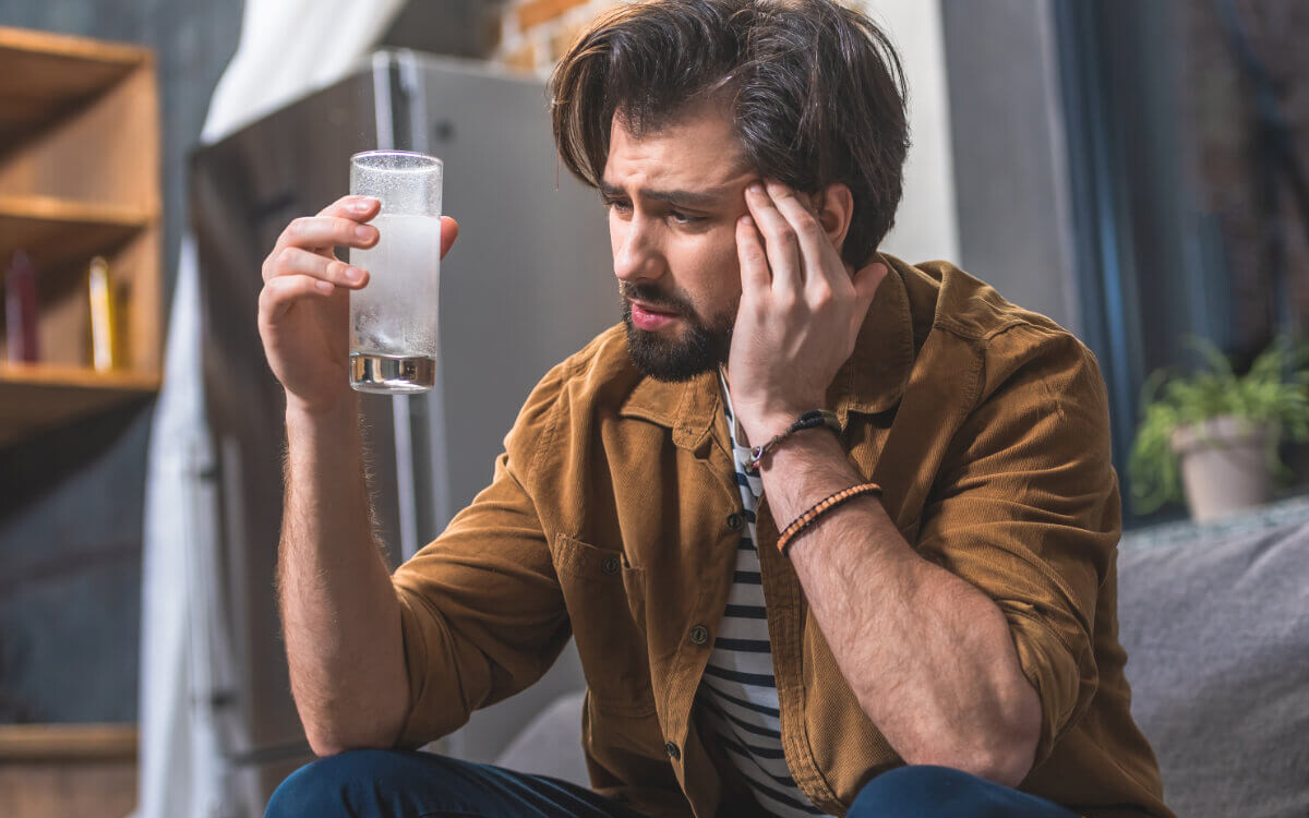 Poppers Nebenwirkungen Symbolbild: Ein Mann hält sich den Kopf vor Schmerzen und hält ein Glas mit Schmerzmittel in der Hand. Englisch: Side effects of poppers Symbolic image: A man is holding his head in pain and holding a glass of painkillers in his hand.