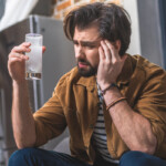 Poppers Nebenwirkungen Symbolbild: Ein Mann hält sich den Kopf vor Schmerzen und hält ein Glas mit Schmerzmittel in der Hand. Englisch: Side effects of poppers Symbolic image: A man is holding his head in pain and holding a glass of painkillers in his hand.