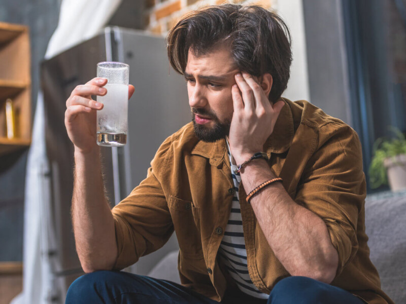 Poppers Nebenwirkungen Symbolbild: Ein Mann hält sich den Kopf vor Schmerzen und hält ein Glas mit Schmerzmittel in der Hand. Englisch: Side effects of poppers Symbolic image: A man is holding his head in pain and holding a glass of painkillers in his hand.
