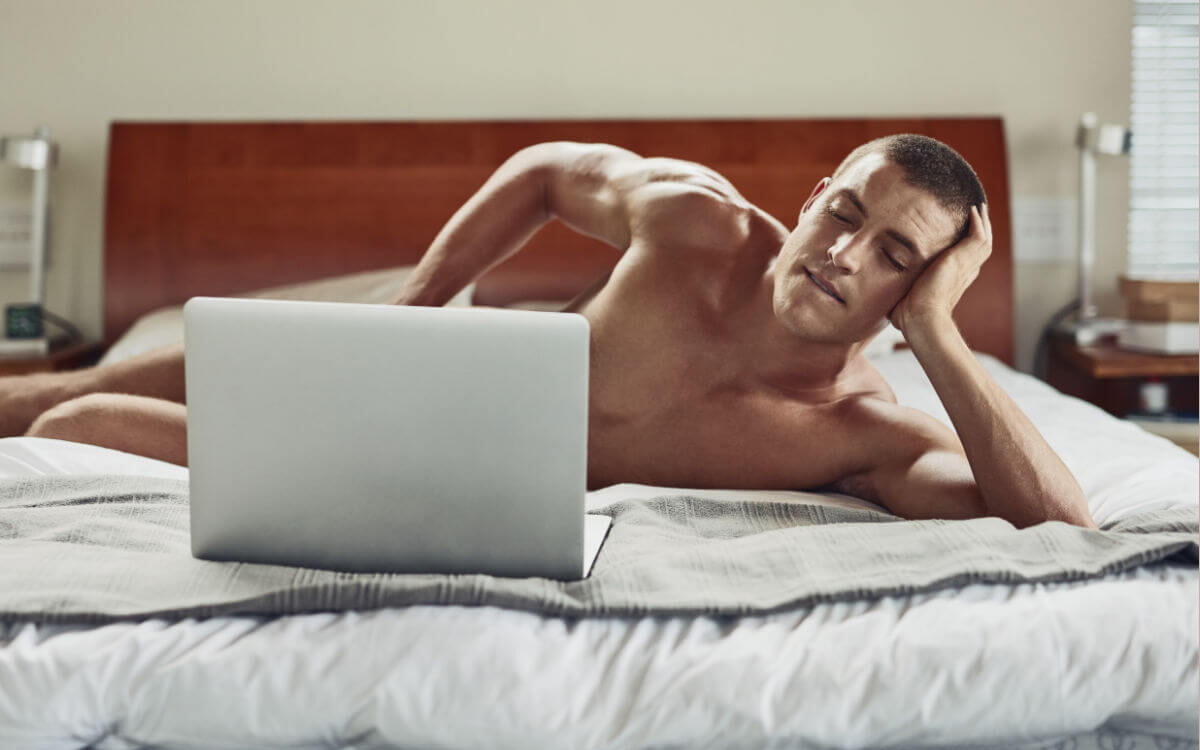 Pornosexuell Symbolbild: Ein Mann liegt nackt auf dem Bett und schaut in einen Laptop, der sein Geschlechtsteil verdeckt. Englisch: Pornosexual symbol: a man lies naked on the bed and looks at a laptop that covers his genitals.