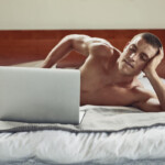 Pornosexuell Symbolbild: Ein Mann liegt nackt auf dem Bett und schaut in einen Laptop, der sein Geschlechtsteil verdeckt. Englisch: Pornosexual symbol: a man lies naked on the bed and looks at a laptop that covers his genitals.