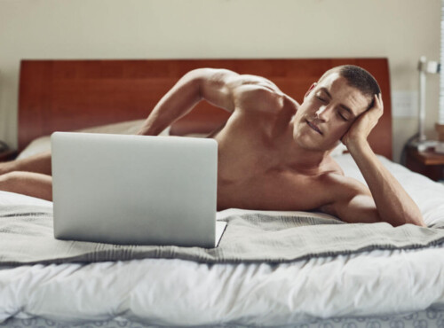 Pornosexuell Symbolbild: Ein Mann liegt nackt auf dem Bett und schaut in einen Laptop, der sein Geschlechtsteil verdeckt. Englisch: Pornosexual symbol: a man lies naked on the bed and looks at a laptop that covers his genitals.