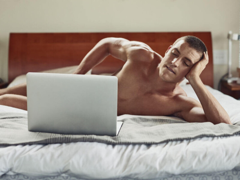 Pornosexuell Symbolbild: Ein Mann liegt nackt auf dem Bett und schaut in einen Laptop, der sein Geschlechtsteil verdeckt. Englisch: Pornosexual symbol: a man lies naked on the bed and looks at a laptop that covers his genitals.