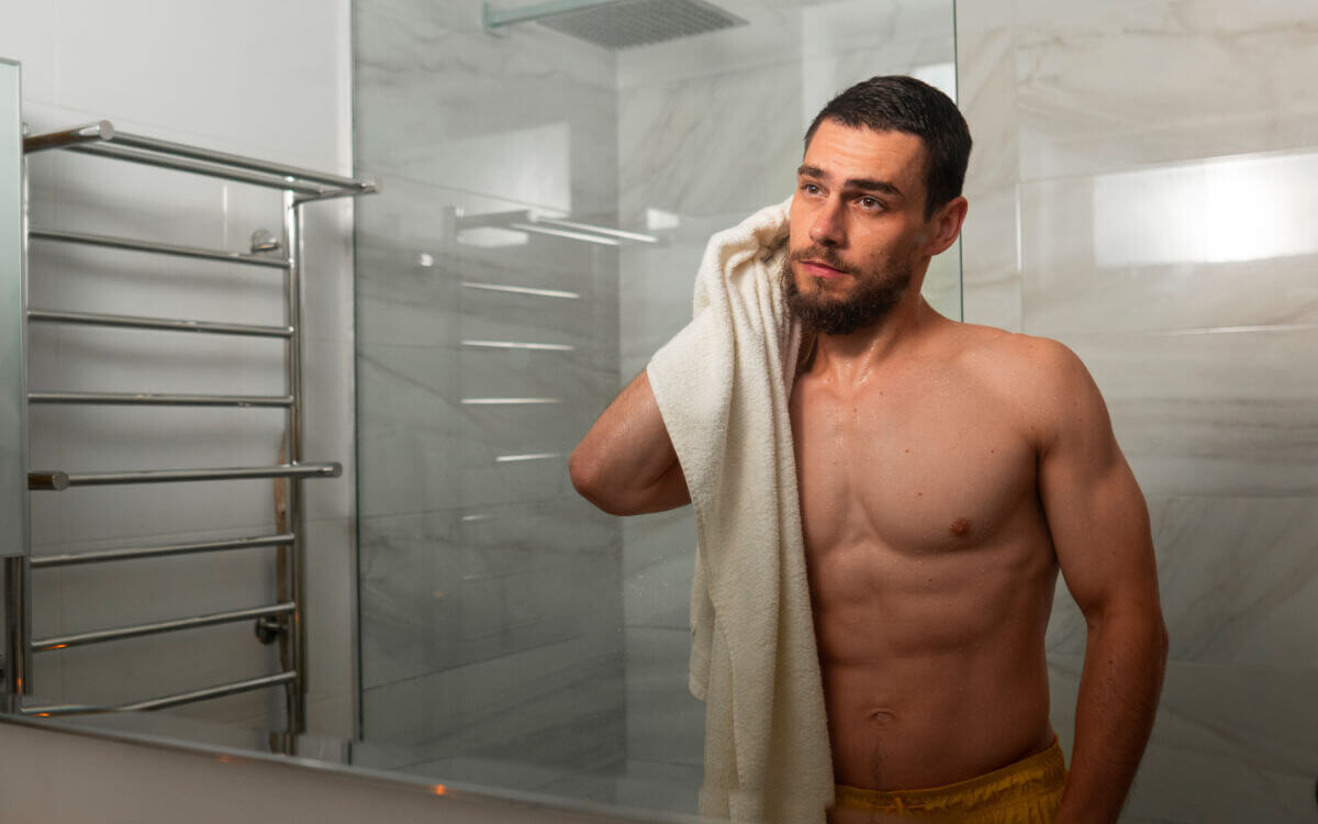 Aftercare für Tops und Doms Symbolbild Topdrop: Ein Mann steht nackt vor dem Badezimmerspiegel und trocknet sich mit einem Handtuch ab. . Aftercare for tops and doms Symbolic image of a top drop: A man stands naked in front of the bathroom mirror and dries himself with a towel.