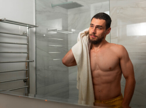 Aftercare für Tops und Doms Symbolbild Topdrop: Ein Mann steht nackt vor dem Badezimmerspiegel und trocknet sich mit einem Handtuch ab. . Aftercare for tops and doms Symbolic image of a top drop: A man stands naked in front of the bathroom mirror and dries himself with a towel.