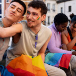 Erfahrungen queerer Menschen in Schule und Ausbildung Symbolbild: Zwei männliche Teenager sowie zwei weibliche sitzen auf einer Bank und halten die Regenbogen-Flagge auf den Beinen. Die beiden Jungs machen ein Selfie. Die Mädchen schauen gemeinsam in ein Handy. . Experiences of queer people in school and education Symbolic image: Two male teenagers and two female teenagers are sitting on a bench holding a rainbow flag between them. The two boys are taking a selfie. The girls are looking at a cell phone together.
