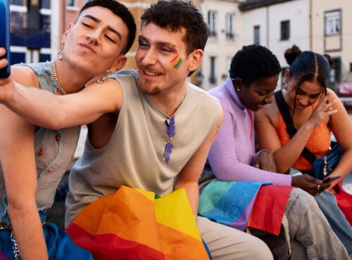 Erfahrungen queerer Menschen in Schule und Ausbildung Symbolbild: Zwei männliche Teenager sowie zwei weibliche sitzen auf einer Bank und halten die Regenbogen-Flagge auf den Beinen. Die beiden Jungs machen ein Selfie. Die Mädchen schauen gemeinsam in ein Handy. . Experiences of queer people in school and education Symbolic image: Two male teenagers and two female teenagers are sitting on a bench holding a rainbow flag between them. The two boys are taking a selfie. The girls are looking at a cell phone together.