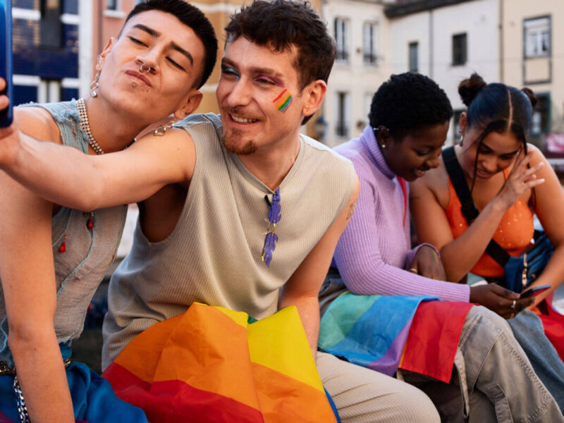 Erfahrungen queerer Menschen in Schule und Ausbildung Symbolbild: Zwei männliche Teenager sowie zwei weibliche sitzen auf einer Bank und halten die Regenbogen-Flagge auf den Beinen. Die beiden Jungs machen ein Selfie. Die Mädchen schauen gemeinsam in ein Handy. . Experiences of queer people in school and education Symbolic image: Two male teenagers and two female teenagers are sitting on a bench holding a rainbow flag between them. The two boys are taking a selfie. The girls are looking at a cell phone together.