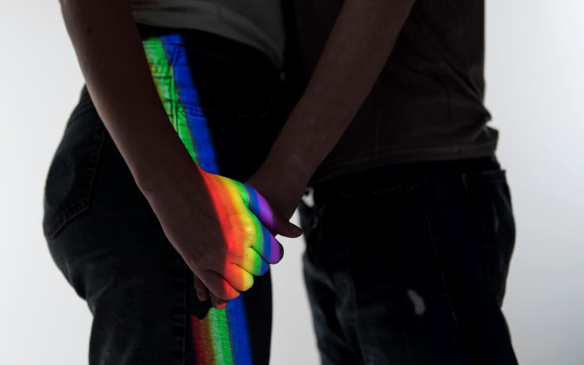 Homophobie Symbolbild: Die Schatten zwei Männer, die sich gegenüberstehend an der Hand halten werden von einem Regenbogen-Licht angestrahlt. Homophobia Symbolic image: The shadows of two men standing opposite each other, holding hands, are illuminated by rainbow light.
