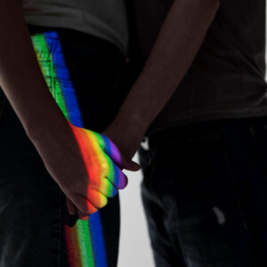 Homophobie Symbolbild: Die Schatten zwei Männer, die sich gegenüberstehend an der Hand halten werden von einem Regenbogen-Licht angestrahlt. Homophobia Symbolic image: The shadows of two men standing opposite each other, holding hands, are illuminated by rainbow light.