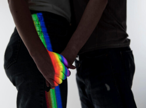 Homophobie Symbolbild: Die Schatten zwei Männer, die sich gegenüberstehend an der Hand halten werden von einem Regenbogen-Licht angestrahlt. Homophobia Symbolic image: The shadows of two men standing opposite each other, holding hands, are illuminated by rainbow light.