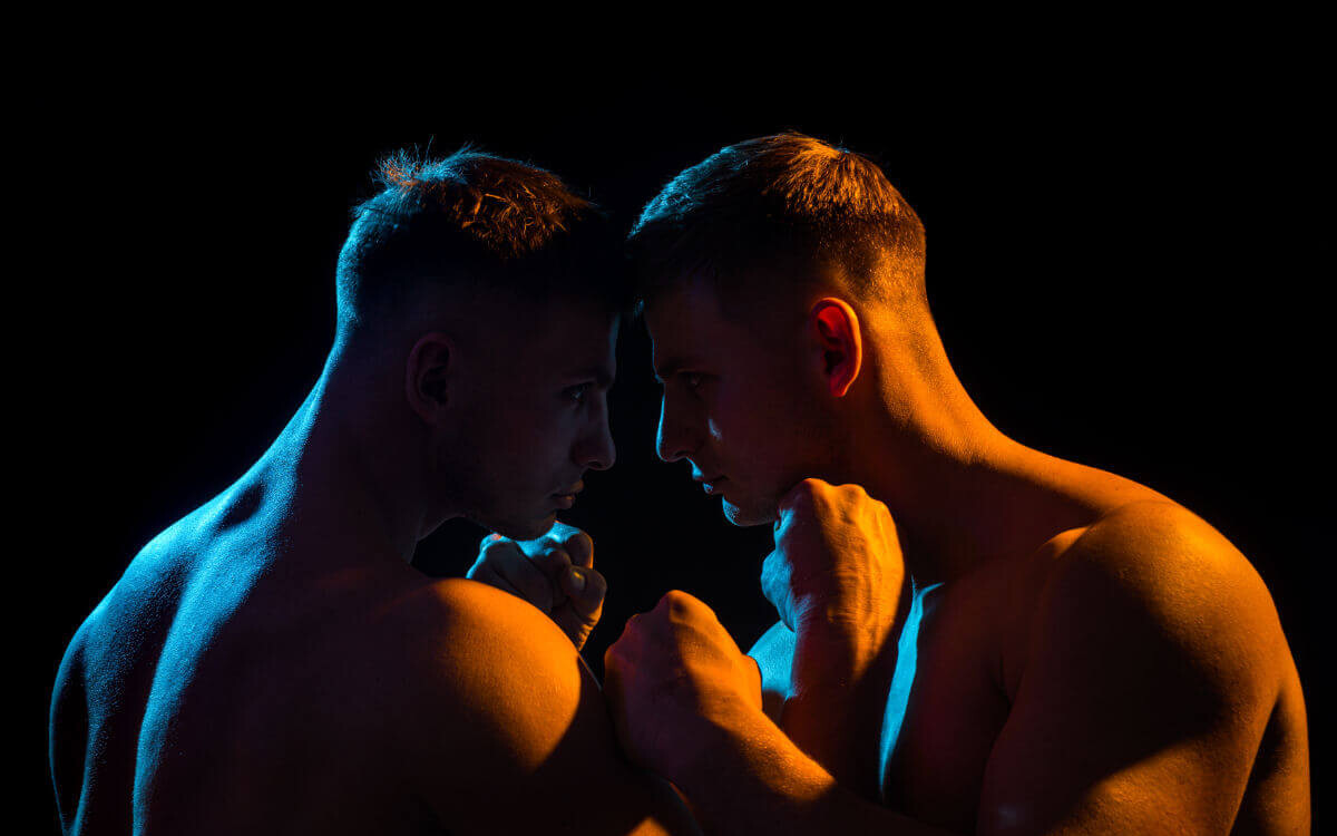 WRAP BDSM Symbolbild: Die nackten Oberkörper zweier Männer, in spärlich orange-farbenes Licht getaucht, die ihre Köpfe aneinanderhalten. . BDSM WRAP Symbolic image: The naked upper bodies of two men, bathed in sparse orange light, holding their heads together.