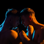 WRAP BDSM Symbolbild: Die nackten Oberkörper zweier Männer, in spärlich orange-farbenes Licht getaucht, die ihre Köpfe aneinanderhalten. . BDSM WRAP Symbolic image: The naked upper bodies of two men, bathed in sparse orange light, holding their heads together.
