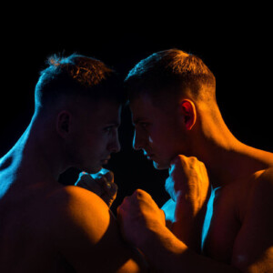 WRAP BDSM Symbolbild: Die nackten Oberkörper zweier Männer, in spärlich orange-farbenes Licht getaucht, die ihre Köpfe aneinanderhalten. . BDSM WRAP Symbolic image: The naked upper bodies of two men, bathed in sparse orange light, holding their heads together.