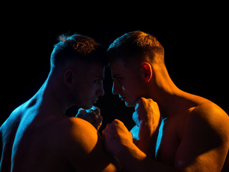 WRAP BDSM Symbolbild: Die nackten Oberkörper zweier Männer, in spärlich orange-farbenes Licht getaucht, die ihre Köpfe aneinanderhalten. . BDSM WRAP Symbolic image: The naked upper bodies of two men, bathed in sparse orange light, holding their heads together.