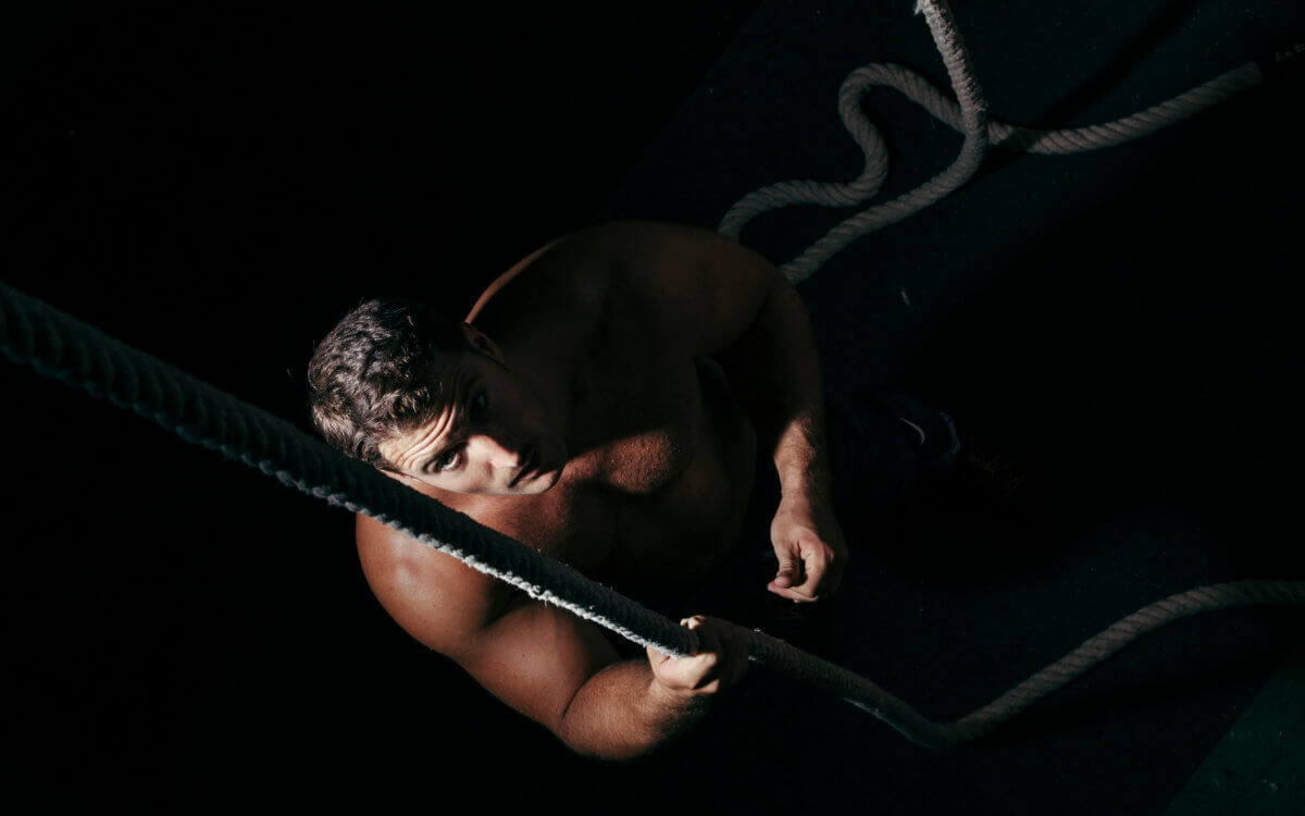 BDSM Shibari Erfahrungsbericht. Symbolbild: Ein Mann sitzt im Dunklen und blickt nach oben in die Kamera. Er ist nackt und mit einem Seil gefesselt. BDSM Shibari experience report. Symbolic image: A man sits in the dark and looks up at the camera. He is naked and tied up with rope.
