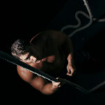 BDSM Shibari Erfahrungsbericht. Symbolbild: Ein Mann sitzt im Dunklen und blickt nach oben in die Kamera. Er ist nackt und mit einem Seil gefesselt. BDSM Shibari experience report. Symbolic image: A man sits in the dark and looks up at the camera. He is naked and tied up with rope.