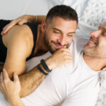 Dry Humping Positionen. Symbolbild: Zwei Männer liegen lachend und sich umarmend auf dem Bett. Englisch: Dry humping positions. Symbolic image: Two men lying on the bed, laughing and hugging each other.