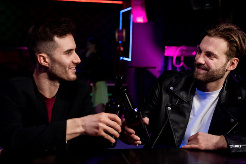 Poppers im Club Symbolbild: Zwei Männer sitzen im Club an der Bar und lächeln sich an. Poppers in a club Symbolic image: Two men sitting at the bar in a club, smiling at each other.