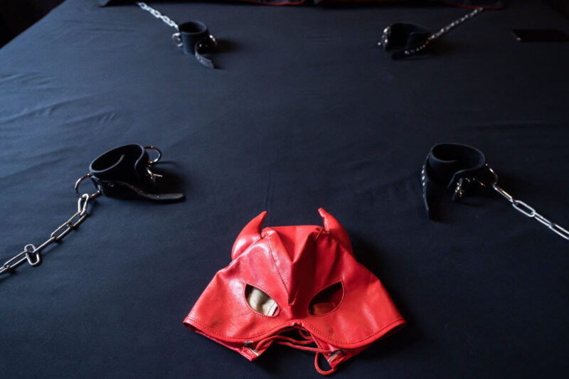 Schwarze Liegefläche mit Fixierungen und roter Leder-Maske als diskrete Szene für einen Dungeon für kleine Wohnungen, stilvoll und platzsparend eingerichtet. Englisch: Black padded surface with restraints and a red leather mask, symbolizing a discreet dungeon for small flats with a stylish, space-saving setup.