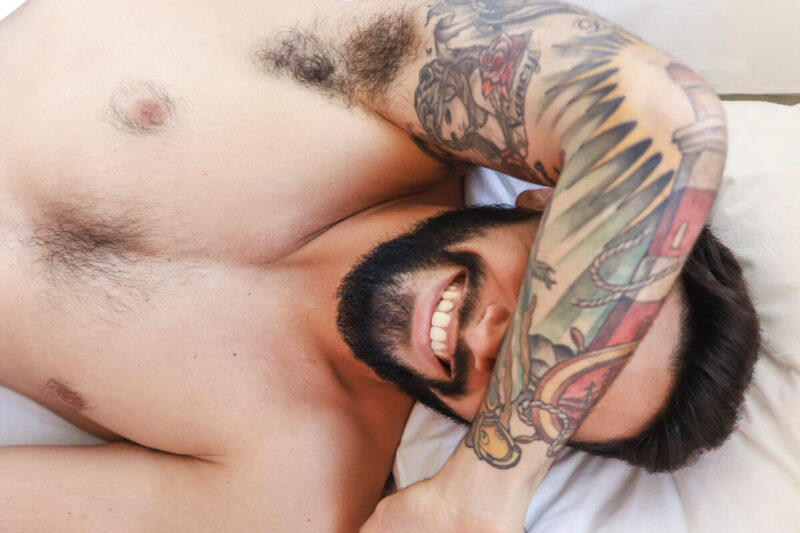 Lächelnder tätowierter Mann liegt entspannt im Bett mit Arm über den Augen, symbolisch für Nähe und Intimität beim gay rollenwechsel. Smiling tattooed man lying relaxed in bed with arm over his eyes, evoking intimacy and connection in a gay role reversal context.
