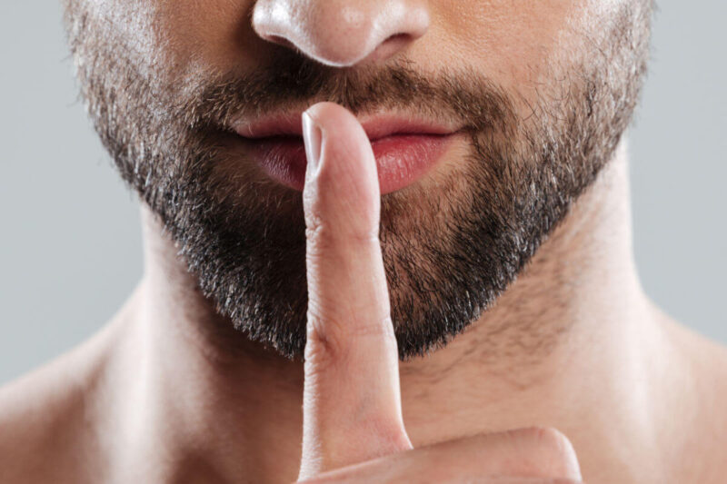 Nahaufnahme eines bärtigen Mannes mit Finger vor den Lippen, Symbol für Lippenpflege für Männer und gepflegte, weiche Lippen im Winter. Englisch: Close-up of a bearded man holding a finger in front of his lips, symbolizing lip care for men and soft, well-groomed lips in winter.