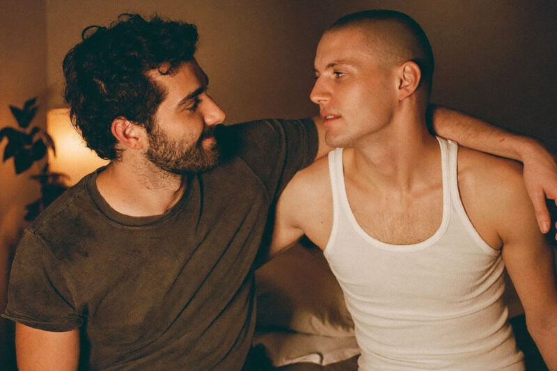 Schwule offene Beziehung Symbolbild: Zwei Männer sitzen eng beieinander auf einem Bett – vertraut, offen und kommunikativ in warmer Lichtstimmung. Two men in a gay open relationship sitting close together on a bed – intimate, communicative and trusting in warm lighting.