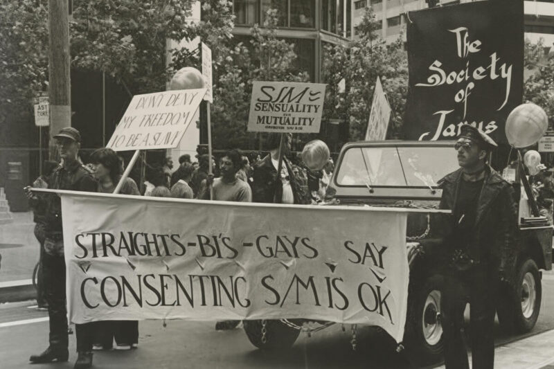 Society of Janus: Historische Aufnahme einer Pride-Parade mit Demonstrierenden und Bannern der Society of Janus, die sich für Konsens, BDSM-Aufklärung und sexuelle Vielfalt einsetzen. English: Society of Janus: Historic photo of a Pride parade featuring Society of Janus banners, highlighting consent, BDSM education, and LGBTQIA+ visibility.