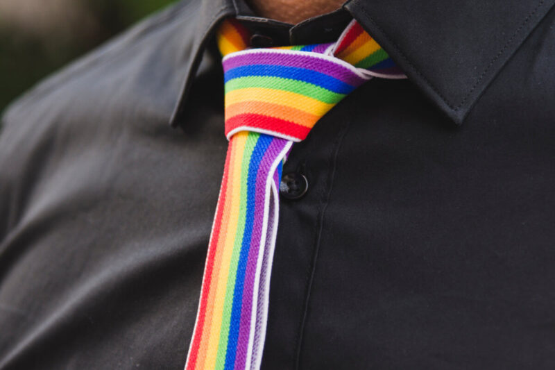 Coming-out am Arbeitsplatz: Person in schwarzem Hemd mit Regenbogenkrawatte als Symbol für LGBTQIA+, Vielfalt und Offenheit im Berufsleben. Coming out at work: Person wearing a black shirt and rainbow tie, symbolizing LGBTQIA+ visibility, diversity and inclusion in the workplace.